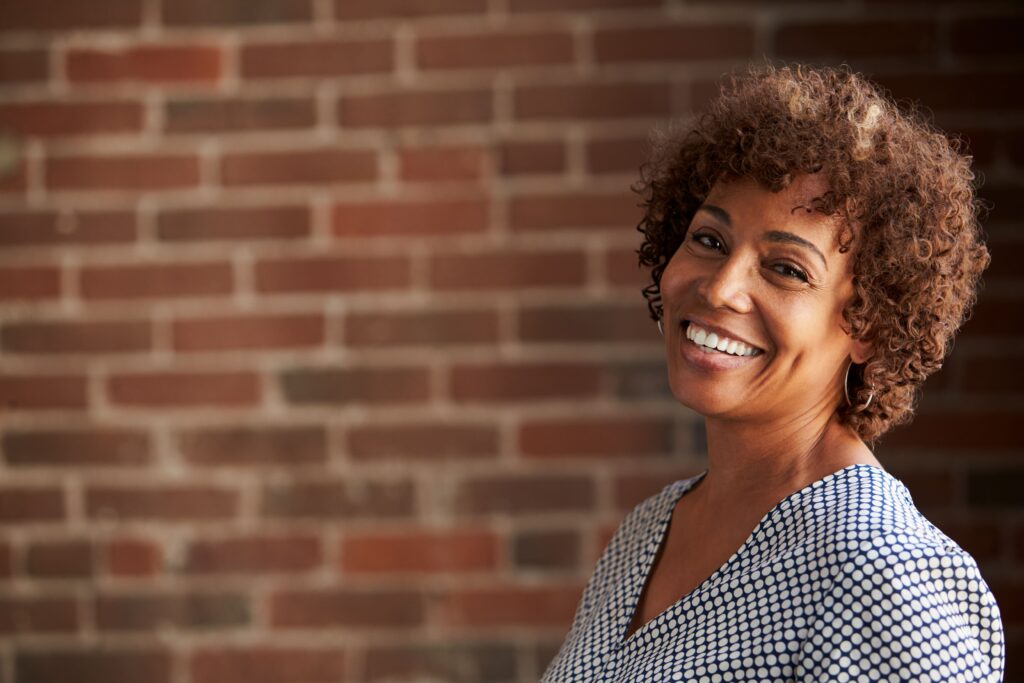 Smiling woman with curly hair in a patterned top stands against a brick wall, conveying warmth and positivity.