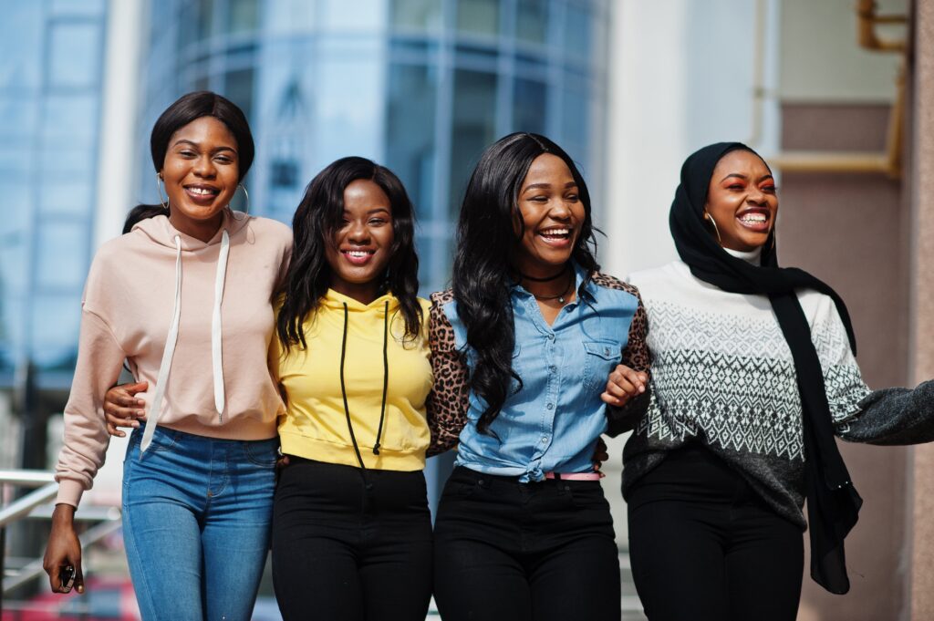 Four women walking arm in arm, smiling joyfully. They wear colorful casual clothes, standing against a modern glass building backdrop, conveying friendship and happiness.