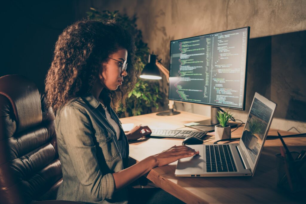 A woman wearing glasses works at a wooden desk with a laptop and dual monitors displaying code. A desk lamp casts a soft light, creating a focused ambiance.