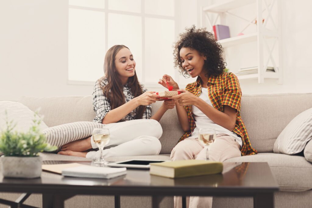Two women sit on a couch in a bright living room, smiling and exchanging a gift. A coffee table with wine glasses and books is in the foreground.