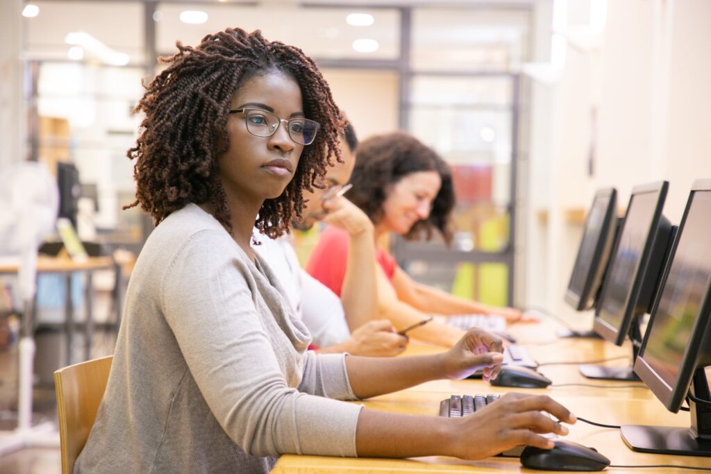 A focused woman with glasses and curly hair works on a computer in a bright classroom. Behind her, others use computers, creating a studious atmosphere.