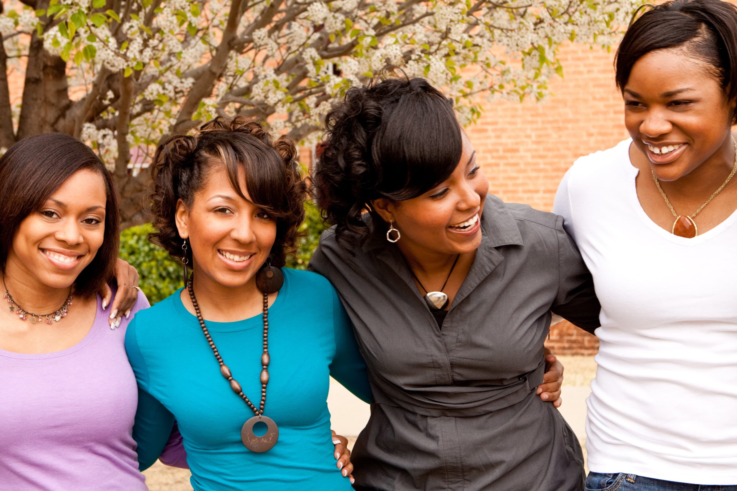 Four women stand together outside, smiling and embracing under a blooming tree. The scene is joyful and warm, conveying friendship and togetherness.