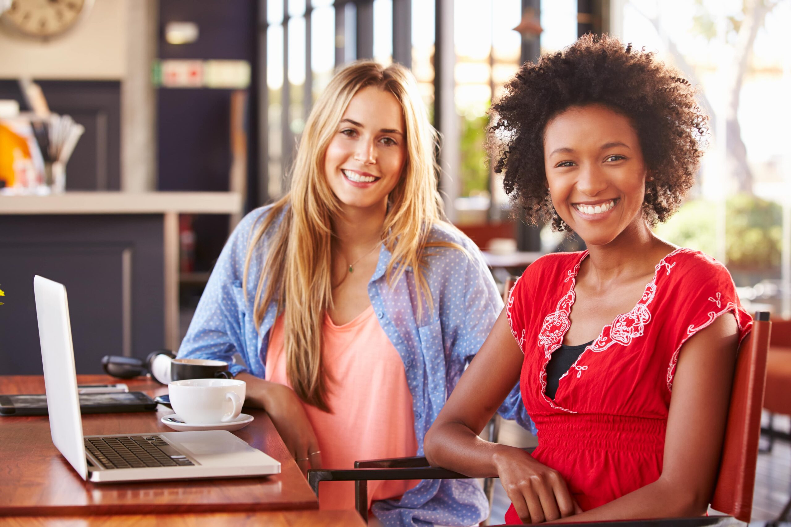 Two women seated at a cafe table smile at the camera. One wears a peach top and blue shirt, the other a red dress. A laptop and coffee cup are on the table, suggesting a casual, friendly meeting.