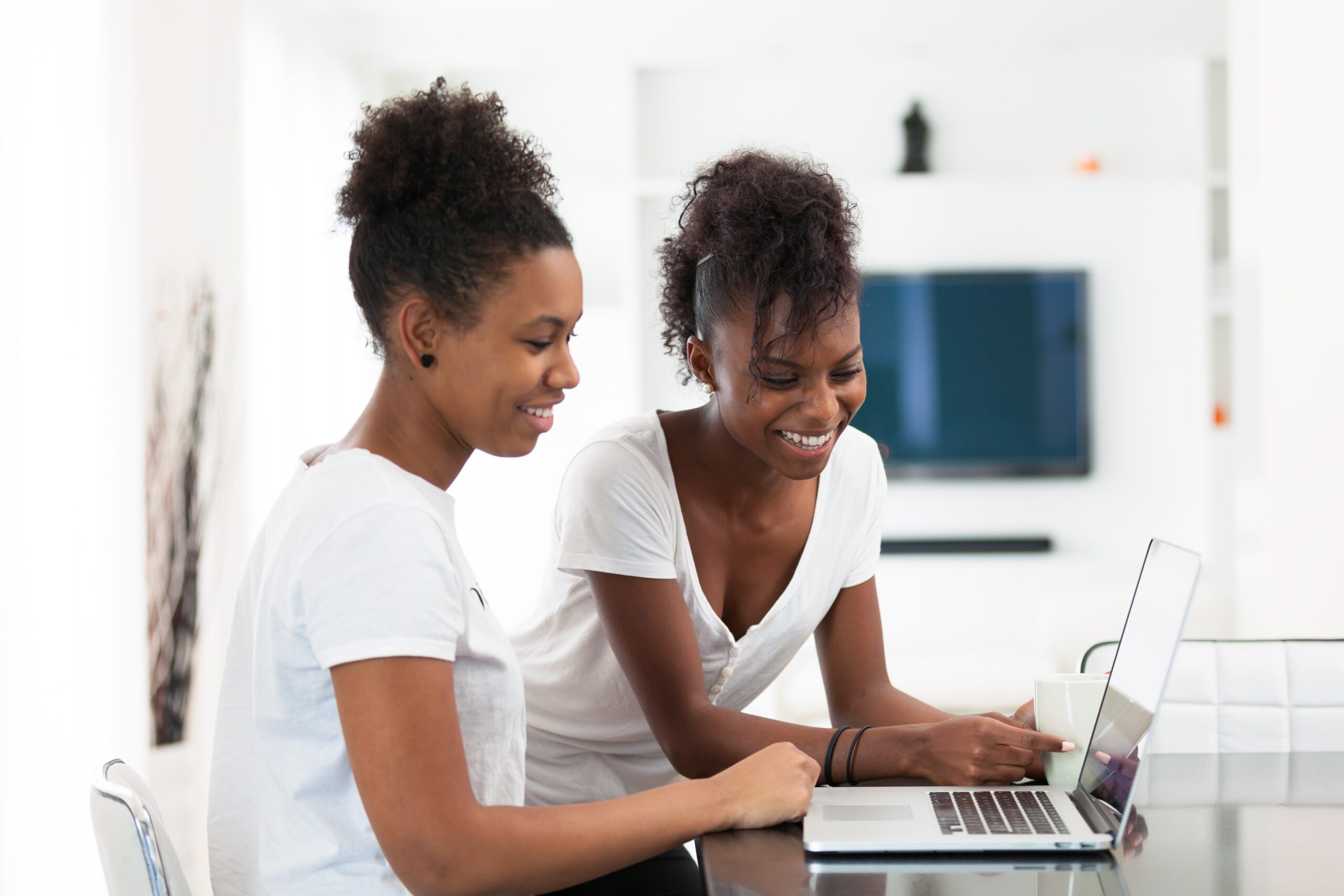 Two women in white shirts happily work on a laptop at a table in a bright, modern living room, conveying a collaborative and cheerful atmosphere.