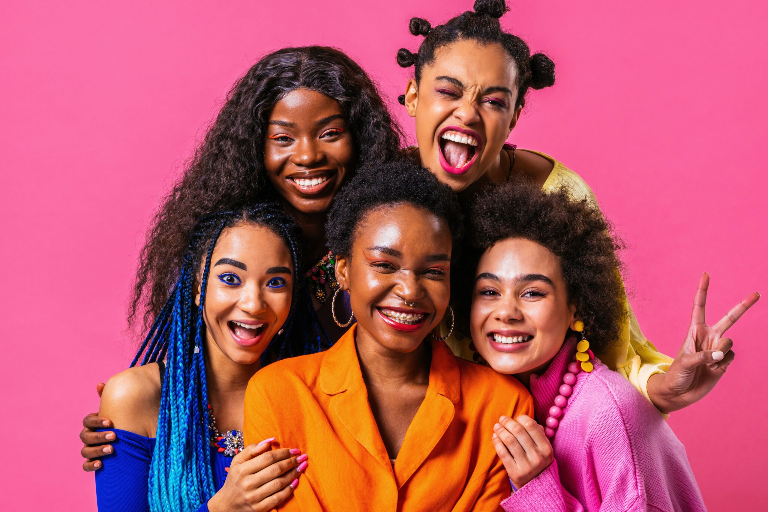 A group of five smiling friends in colorful outfits and diverse hairstyles pose against a bright pink background, exuding joy and camaraderie.