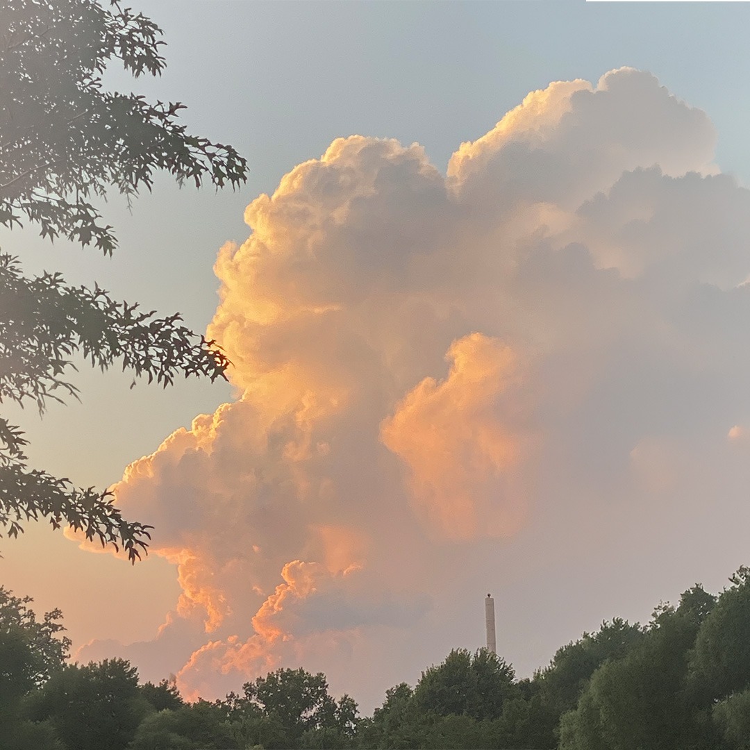 Dramatic clouds glow orange against a twilight sky, framed by trees, with a distant smokestack visible, creating a serene yet industrial landscape.