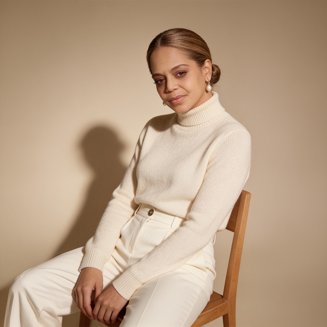 A woman sits gracefully on a wooden chair, wearing a cozy cream turtleneck and white trousers, set against a soft beige background.